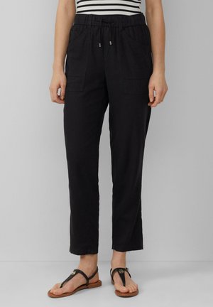 Vrouw draagt zwarte broek met trekkoord en voorzakken, gestreept bovenstuk en zwarte platte sandalen, staand tegen een effen achtergrond.