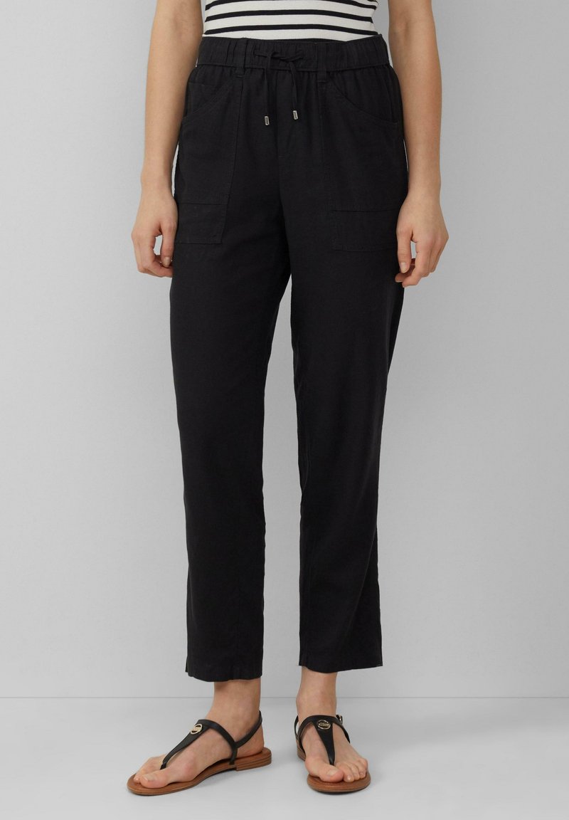 Femme portant un pantalon noir à taille coulissante avec poches devant, un haut rayé et des sandales plates noires, debout devant un fond uni.