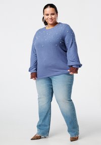 Vrouw in een blauwe trui met pareldetails, bruine kanten manchetten, lichtblauwe spijkerbroek, luipaardprint hakken en grote oorbellen, staan en glimlachen.