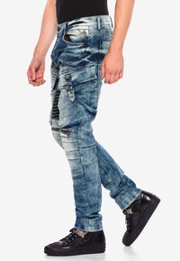 Denim cargobroek in een lichtblauwe, gewassen deterioratie, met meerdere zakken, ritsen en verstelbare banden; gecombineerd met zwarte sneakers.