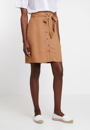Femme portant une jupe marron boutonnée à mi-cuisse avec une ceinture nouée à la taille, un haut blanc et des espadrilles blanches, debout sur un fond uni.