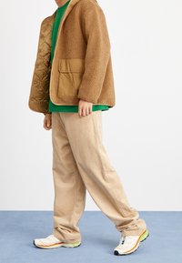 Chaqueta marrón tipo sherpa con interior acolchado y bolsillos de parche en el frente, sobre una camiseta verde. Pantalones de pana beige y zapatillas de correr multicolores.