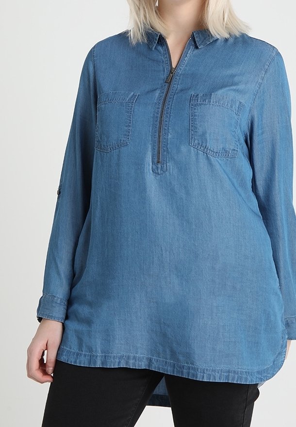 Chemise en denim à manches longues ; elle possède une fermeture éclair quart-de-tour, deux poches poitrine, et un ourlet allongé. Le tissu est bleu moyen et légèrement texturé.