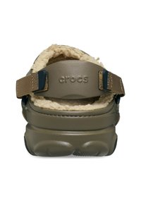 Crocs de color verde oliva con forro beige acogedor y correa marrón ajustable. Presentan una suela texturizada y forma redondeada para mayor comodidad.