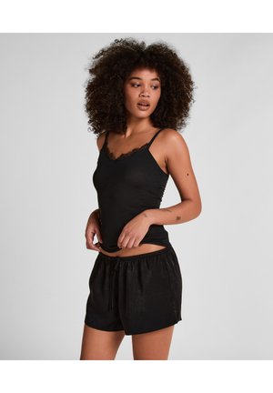 Vrouw met krullend haar, gekleed in een zwart hemdje met kanten rand en zwarte losse korte broek, tegen een effen grijze achtergrond.