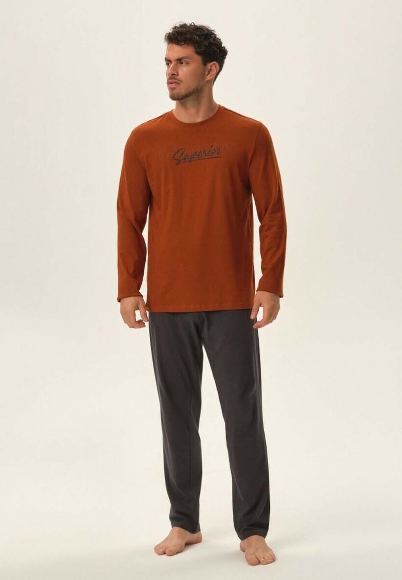 Męski zestaw odzieży do relaksu, składający się z długiego rękawa w kolorze wypalonej pomarańczy z czarnym logo oraz ciemnoszarych, luźnych spodni. Gładka faktura materiału.