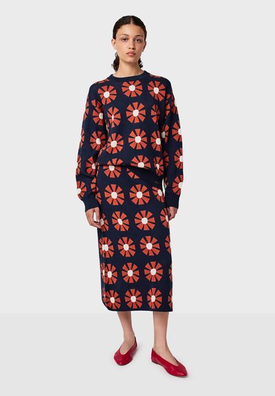 Stickad tröja och kjol i marinblått med röd och vit blommönster. Långa ärmar, rundad halsringning och knä-lång kjol. Röda ballerina skor.