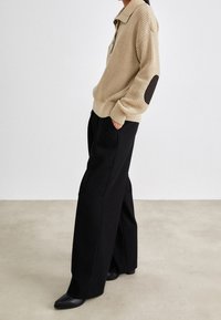 Persoon met een beige gestructureerde trui met zwarte elleboogpatches, zwarte wijde broek en zwarte schoenen, staand met de handen in de zakken.