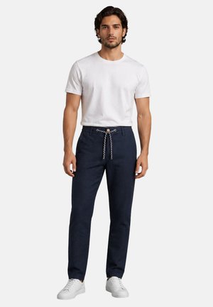Man wearing white short-sleeve t-shirt, navy blue pants with black and white rope belt, and white sneakers, standing against white background.