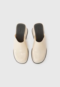 Mule-uri slip-on bej cu un finisaj neted și lucios, vârf pătrat și toc jos. Talpa interioară și exteriorul negre oferă contrast și stabilitate.