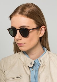 Sunglasses dengan corak karet dengan lensa bulat, lensa gelap, dan lengan ramping. Model tersebut mengenakan jaket kulit ringan dan kemeja biru.
