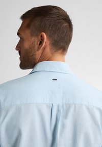 Světle modrá košile s krátkým rukávem a límcem, s malým černým logem vzadu poblíž výstřihu a hladkým, strukturovaným materiálem.