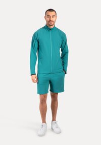 Chaqueta deportiva verde con cuello alto y cierre de cremallera, combinada con pantalones cortos a juego. La tela suave y unas zapatillas blancas completan el conjunto.