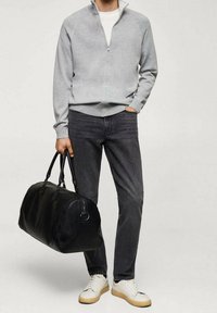 Uomo che indossa un maglione grigio con zip, jeans scuri, sneaker bianche, mentre tiene una borsa da viaggio in pelle nera con la mano sinistra e ha la mano destra nella tasca.