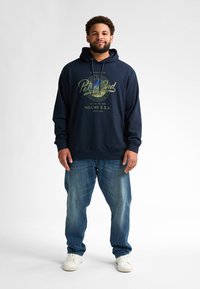 Námořnická modrá mikina s kapucí a zeleným grafickým potiskem, s tkaničkou na kapuci a klokaní kapsou, kombinovaná se světle modrými džíny a bílými teniskami.