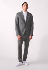Grå teksturert dressjakke med matchende bukser, hvit skjorte med krage, og hvite sneakers. To-knapps frontlukking og sidelommer.