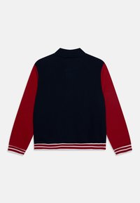 Giacca varsity blu navy e rossa con colletto a costine, maniche rosse e orli rossi con strisce bianche, vista da dietro.