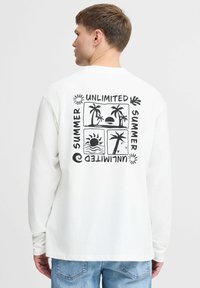 Camiseta blanca de manga larga con diseño gráfico negro que presenta palmeras y motivos del sol, el texto dice "VERANO ILIMITADO", corte relajado.