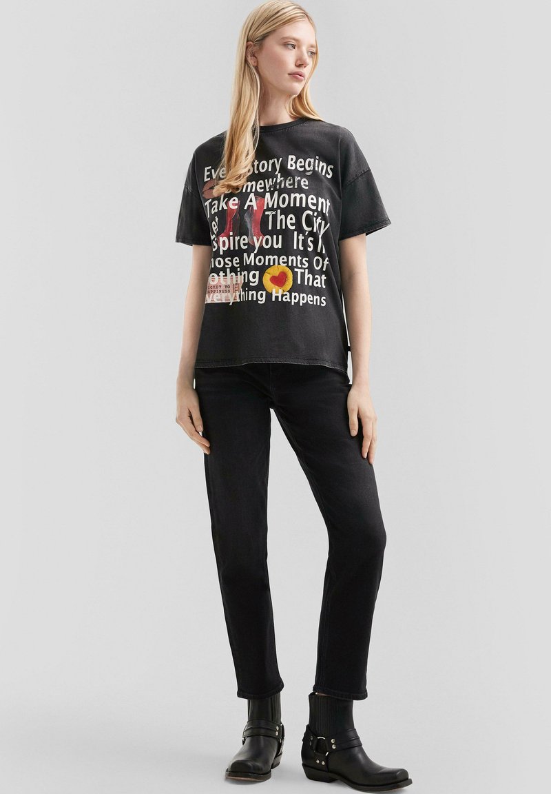 Camiseta de algodón negra con texto gráfico en varios colores. Combinada con jeans negros y botas hasta el tobillo con detalles de hebilla.