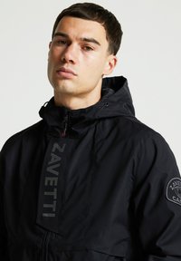 Fekete kapucnis dzseki zipzárral, fényvisszaverő "ZAVETTI" felirattal és kerek logóval az ujjon. Simább anyagtextúra.