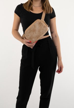 Vrouw in zwart tenue draagt een beige crossbodytas met een getextureerde voorkant en verstelbare riem over haar borst.