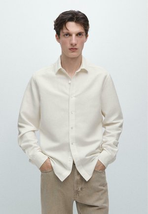 Giovane uomo con capelli corti e scuri, indossa una camicia bianca a maniche lunghe abbottonata e pantaloni beige, in piedi con le mani nelle tasche.