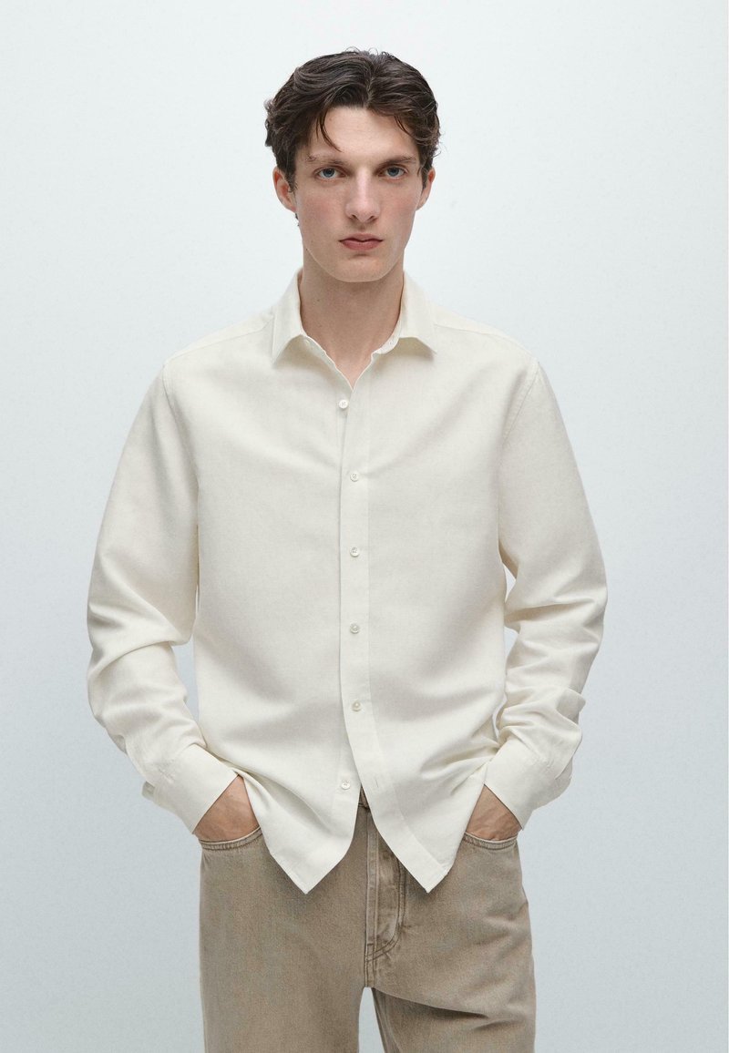 Młody mężczyzna z krótkimi, ciemnymi włosami, ubrany w białą, zapinaną na guziki, długą koszulę oraz beżowe spodnie, stoi z rękami w kieszeniach.