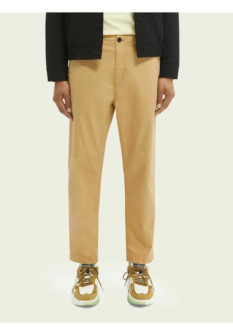 Beige Chinos mit einem geraden Schnitt, aus Baumwolle, mit einem Frontknopfverschluss und Seitentaschen, kombiniert mit braunen und weißen Sneakers.