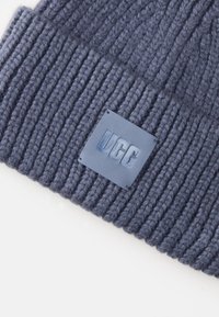 Bonnet en tricot bleu foncé avec une texture côtelée. Présente une étiquette en cuir rectangulaire embossée avec "UGG."