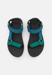 Teva sandale s crnom gumenom potplatom, plavim i zelenim platnenim trakama te podesivom opremom. Imaju ravni dizajn i podstavljenu unutrašnjost.