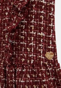 Burgundowa tkanina tweedowa z białymi i złotymi akcentami, z falbaną na krawędzi i przymocowanym złotym logo w kształcie serca.