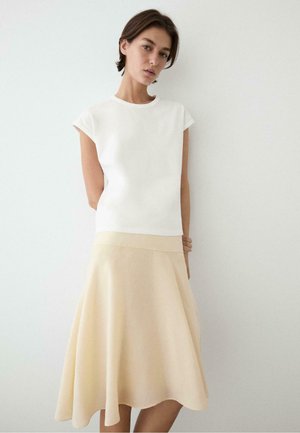 Młoda kobieta w białej bluzce z krótkim rękawem i beżowej spódnicy w kształcie litery A stoi przy gładkiej białej ścianie, patrząc lekko w dół.