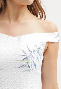 Robe blanche à épaules dénudées en tissu texturé, avec un imprimé floral bleu et vert sur le côté gauche. Détail de fermeture éclair apparente visible.