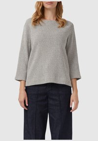 Vrouw in een lichtgrijze gebreide trui met structuur, met driekwart mouwen en een donker hoge taille met wijde benen.