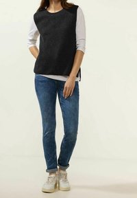 Top noir sans manches en tricot porté sur une chemise blanche à manches longues, associé à un jean bleu et des baskets beige avec des détails texturés.