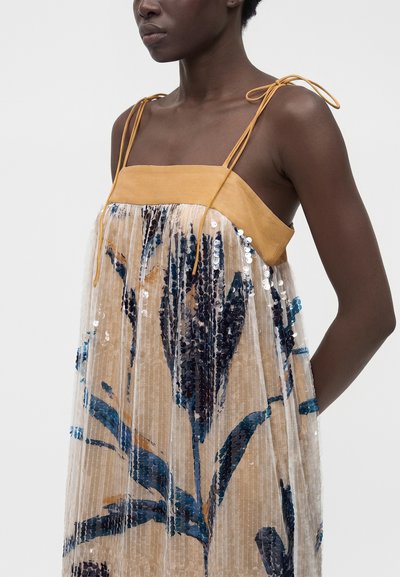 Femme portant une robe sans manches avec des bretelles jaune moutarde et une superposition transparente ornée de motifs floraux en sequins bleus.