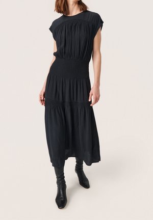 Kobieta ubrana w bezrękawnikową czarną sukienkę midi z marszczeniami oraz czarne botki na obcasie do połowy łydki, stojąca na neutralnym tle.