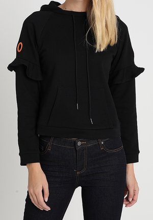 Femme portant un sweat à capuche noir avec des manches à volants et un patch circulaire orange sur le haut du bras gauche, associé à un jean foncé.