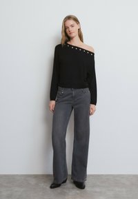 Joven de pie junto a una pared, vistiendo un suéter negro de hombros descubiertos con ojales plateados, jeans grises de pierna ancha y botines negros.