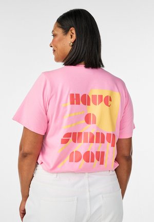 Frau mit schulterlangen schwarzen Haaren, die ein pinkes T-Shirt mit der Aufschrift "Have a sunny day" und einer gelben Sonne als Grafik trägt, kombiniert mit weißen Hosen.