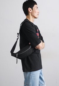 Black fanny pack made of textured material, featuring a bold logo and adjustable strap. Worn crossbody with a sleek, modern design.