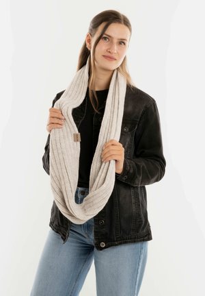 Jonge vrouw in lichtblauwe jeans en een zwarte jas, die een dikke beige gebreide sjaal om haar nek houdt tegen een effen achtergrond.