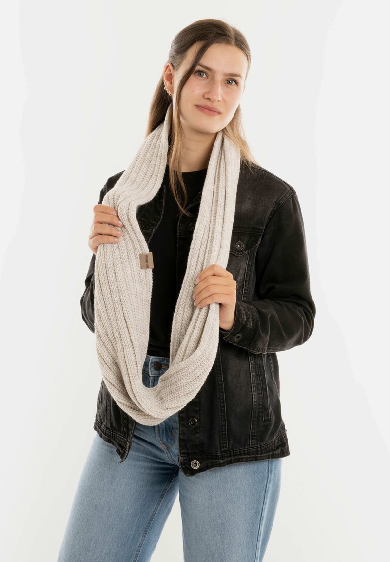 Jonge vrouw in lichtblauwe jeans en een zwarte jas, die een dikke beige gebreide sjaal om haar nek houdt tegen een effen achtergrond.