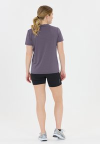 Kvinna med blont hästsvans som bär en lila t-shirt, svarta shorts och grå löparskor, stående med ryggen mot på en vit bakgrund.