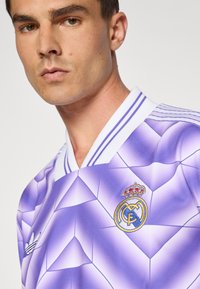 Camisa de fútbol morada con un patrón geométrico, con ribetes blancos y el logo del Real Madrid en el pecho, fabricada con un tejido transpirable.