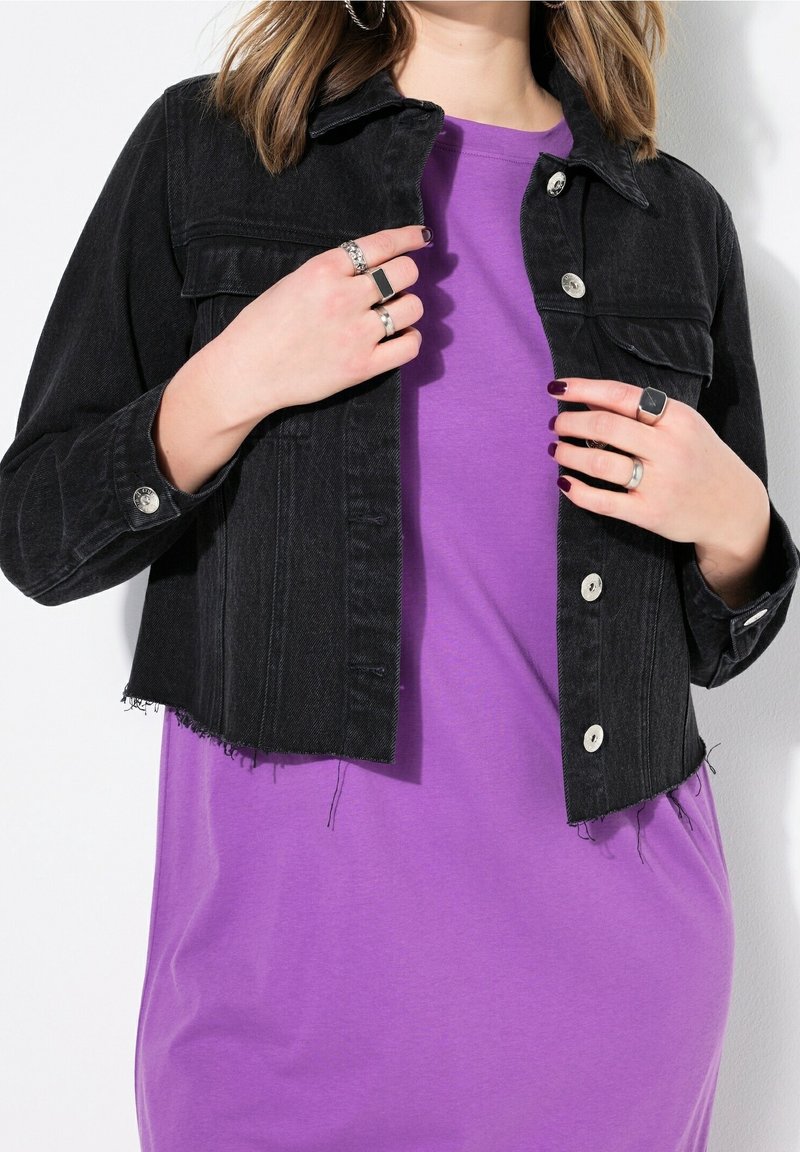 Veste en denim noire courte avec des boutons argentés portée sur un t-shirt violet uni, présentant un ourlet effiloché et une coupe décontractée. Bagues aux doigts.
