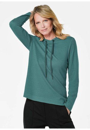 Haut long en teal avec capuche et cordons de serrage, présentant une texture côtelée. Assorti à un pantalon noir, présenté sur fond blanc.