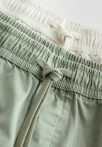 Tailles élastiques avec cordons noués sur des pantalons décontractés vert clair et blanc, montrant la texture du tissu et les détails de couture.