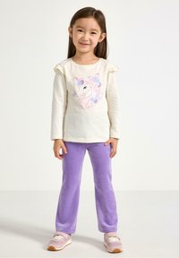 Light cream long-sleeve shirt with ruffled sleeves and unicorn graphic, paired with soft purple pants and light pink sneakers.