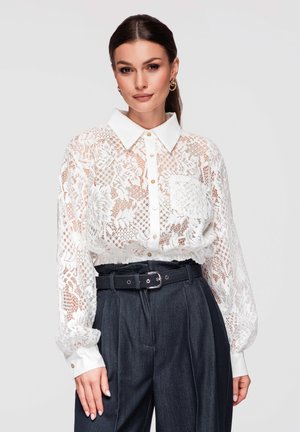 Nainen valkoisessa pitsisessa napitettavassa paitapuserossa, jossa on pitkät hihat, ja tummissa korkeavyötäröisissä housuissa, joissa musta vyö, yksinkertaista taustaa vasten.